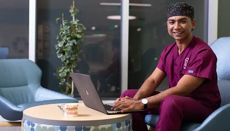 Student wearing scrubs sitting in lobby using laptop